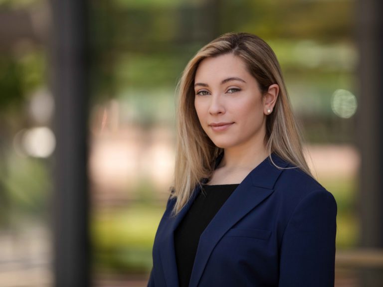 Professional headshot of a young blonde woman wearing a blue blazer and black top, photographed against a soft, blurred green background