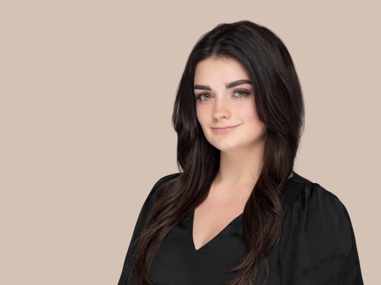 Professional headshot of a young adult woman wearing a black top, photographed against a light tan background